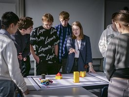 Auf dem Bild steht eine Gruppe von Jugendlichen um eine junge Frau mit schultlangen blonden Haaren. Die Frau deutet mit der rechten Hand auf einen Tisch auf dem ein großes Plakat, ein Legorover und mehrere Leoghindernisse liegen. 